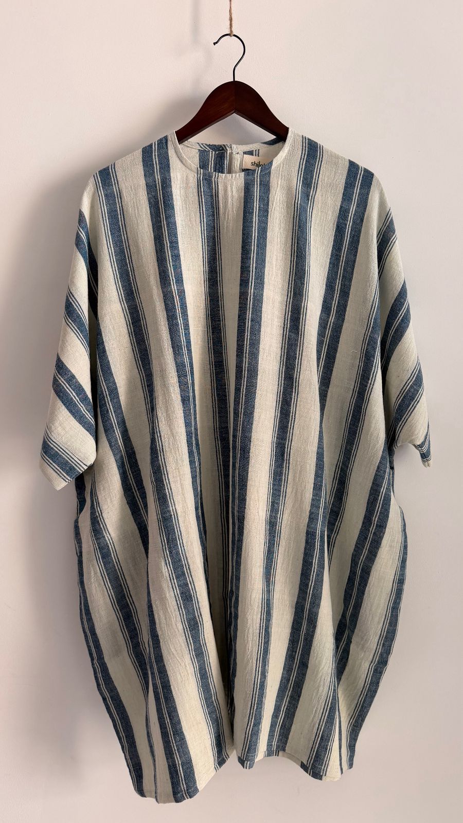 Handwoven cotton Cedar dress in oat with indigo vertical stripes hanging on wooden hanger.